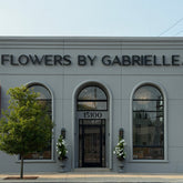 A grey building with three large arched windows and a black letter sign reading "FLOWERS BY GABRIELLE". A tree sits on the left side of the building, and two large planters with mandavilla trees frame the front door.
