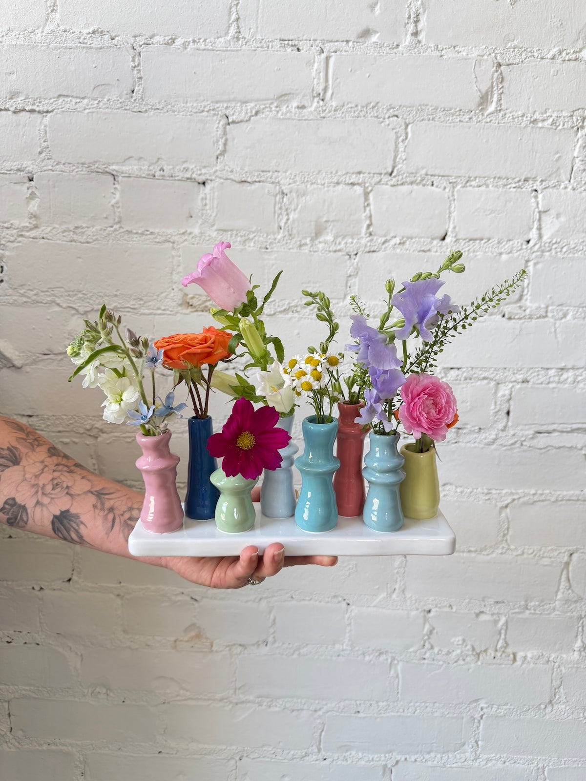 Eight colorful budvases set on a ceramic base, filled with colorful flowers in a wildflower style, against a white brick background. 