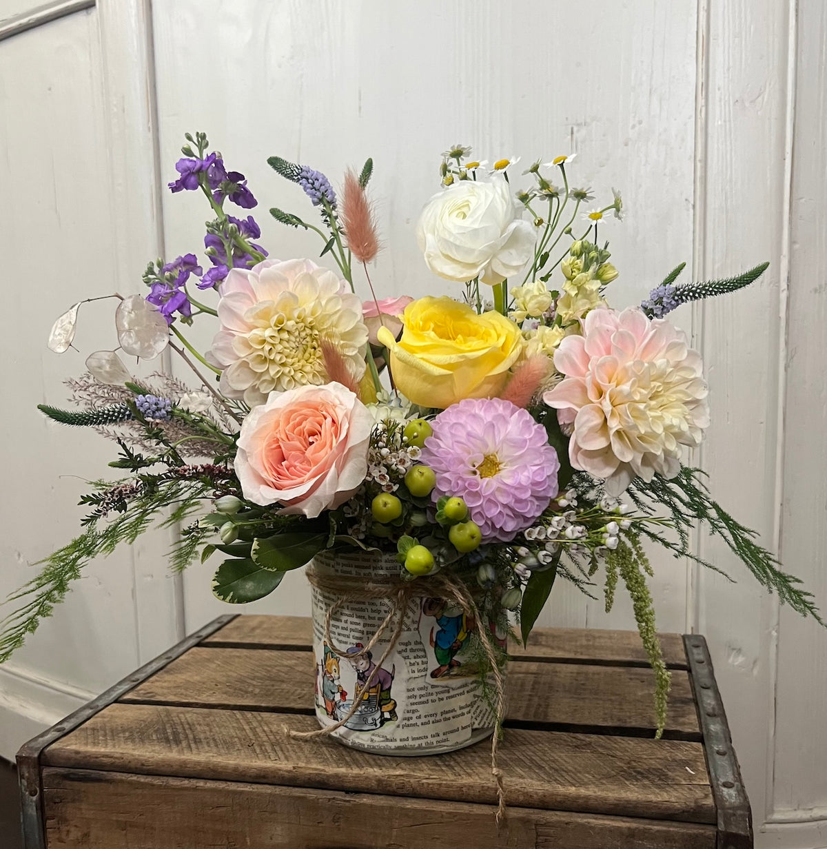 Floral arrangement in soft neutral colors, in a vase decoupaged with pages from a children's book. Arrangement sits atop a wooden crate against a white background.