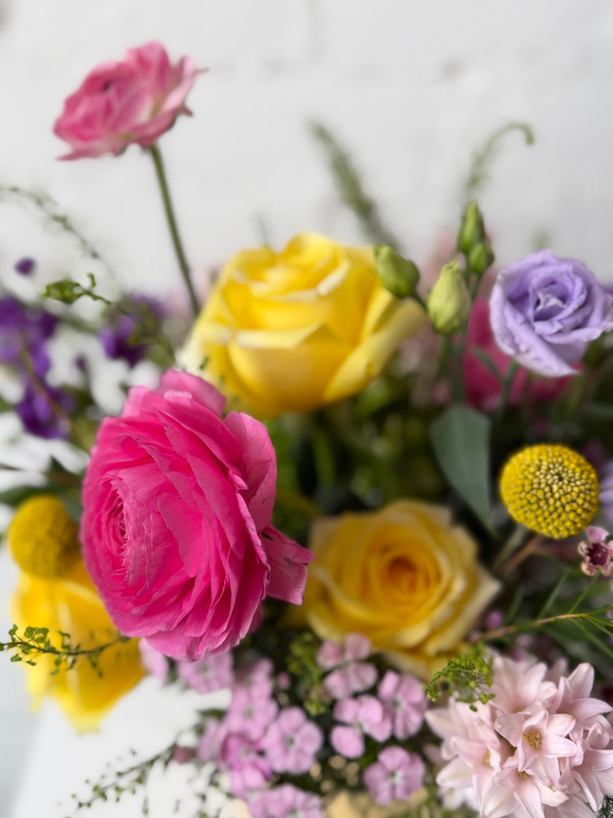 A close-up of a vibrant floral arrangemen featuring bright pink, yellow, and purple blooms with green foliage, set against a white brick wall background.