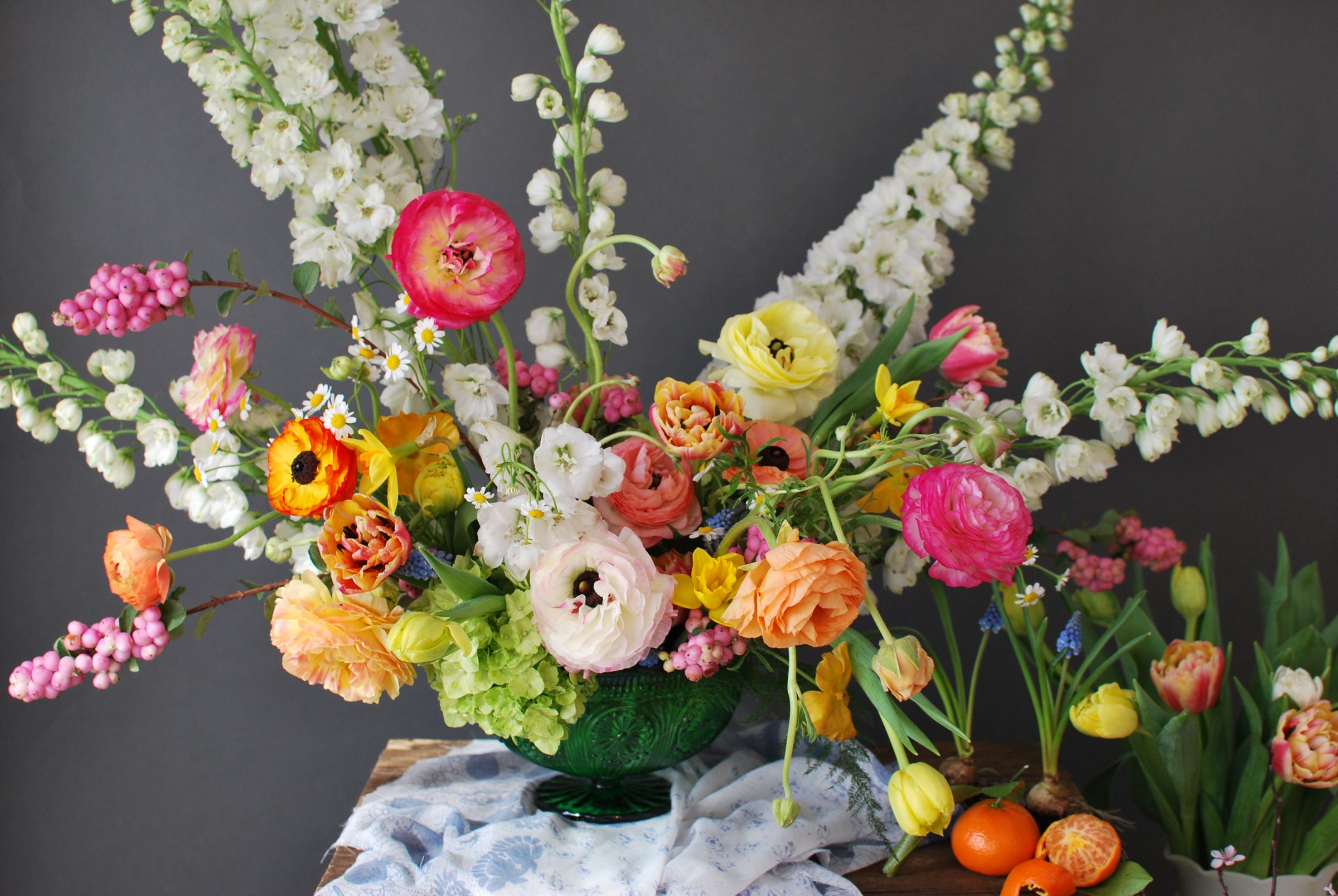 A vibrant, multicolored floral arrangement featuring ranunculus, tulips, hydrangea, and other spring florals in a green glass compote, set against a dark grey background.