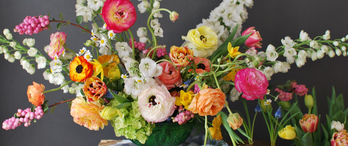 A close up of a vibrant, multicolored floral arrangement featuring ranunculus, tulips, hydrangea, and other spring florals in a green glass compote, set against a dark grey background.