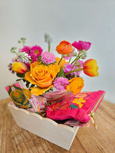A gift box with a bright pink, orange and purple floral arrangement, a pair of pink socks, and a hot pink zip pouch on top of a wooden table. 