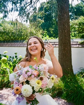 Candid shot of a bride holding her bouquet of pastel flowers, in front of lush green plants and a tree.