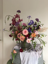 A large, dark wildflower arrangement in a dark vase, set against a plain beige wall with a light gray draped cloth underneath.