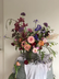A large, dark wildflower arrangement in a dark vase, set against a plain beige wall with a light gray draped cloth underneath.
