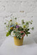 Floral arrangement in soft, neutral colors placed in a light yellow ceramic pot sits against a white brick background. 