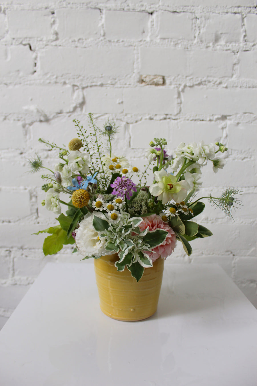 Floral arrangement in soft, neutral colors placed in a light yellow ceramic pot sits against a white brick background. 