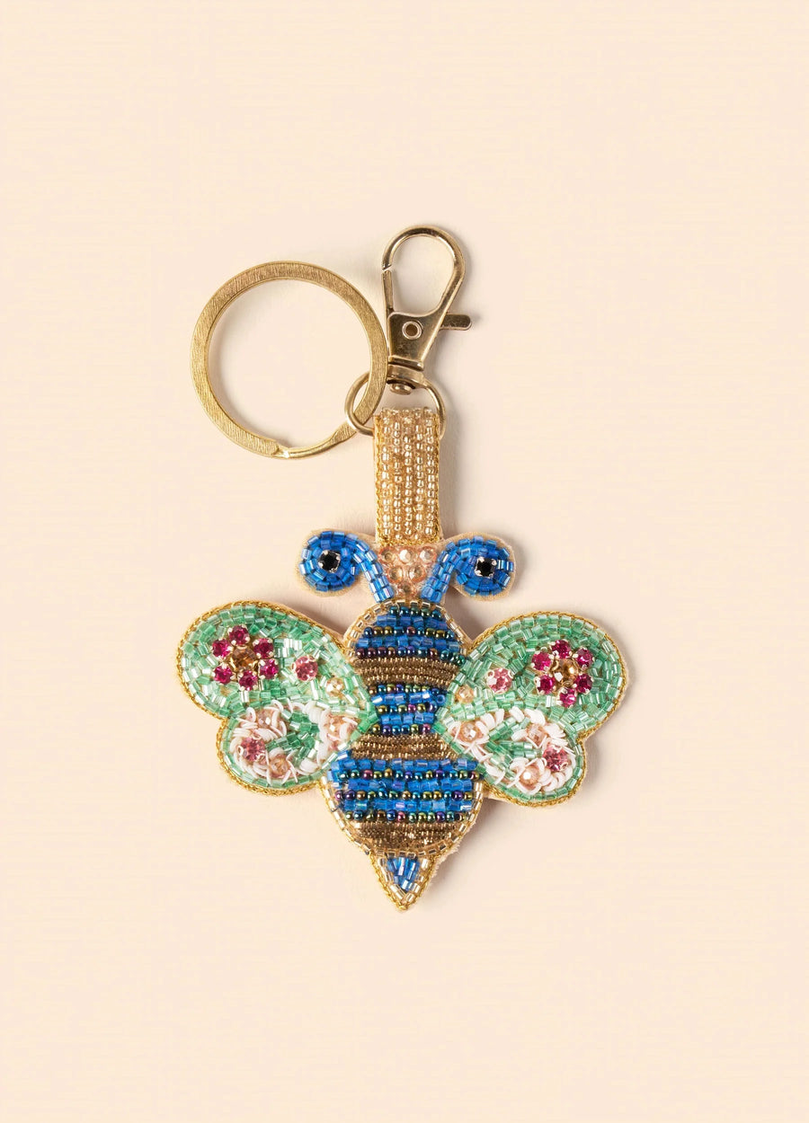 Gold keyring with a beaded blue and mint green bee charm, set against a cream background.