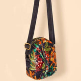 A small crossbody bag with a dark navy floral print, featuring a wide black shoulder strap with a gold buckle, set against a solid beige background.