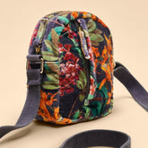 A small crossbody bag with a dark navy blue base and a vibrant multicolor floral print in orange, green, and red. It features a matching dark navy webbed strap and gold-tone hardware, set against a solid beige background.