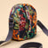 A small crossbody bag with a dark navy blue base and a vibrant multicolor floral print in orange, green, and red. It features a matching dark navy webbed strap and gold-tone hardware, set against a solid beige background.