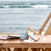 A light blue tin candle sits on a wooden table with vanilla beans in front of a light blue water background.