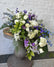 A large casket spray in white, blue, green, and purple flowers, displayed on a wooden table against a white brick wall.