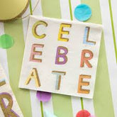 Cocktail napkin with "CELEBRATE" spelled out in colorful block letters, placed on a green and white striped background with confetti.