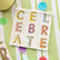 Cocktail napkin with "CELEBRATE" spelled out in colorful block letters, placed on a green and white striped background with confetti.