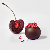 Half bitten cherry next to a chocolate covered cherry, in front of a white background. 
