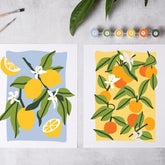 Two colorful fruit paintings displayed side by side on a light gray surface, with paintbrushes and small paint tubes nearby. The left print shows yellow lemons with green leaves on a light blue background, and the right print shows orange citrus fruits with green leaves on a pale yellow background.