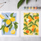 Two colorful fruit paintings displayed side by side on a light gray surface, with paintbrushes and small paint tubes nearby. The left print shows yellow lemons with green leaves on a light blue background, and the right print shows orange citrus fruits with green leaves on a pale yellow background.