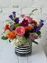 Floral arrangement in bright colors set in a glass vase with a black and white striped ribbon wrapped around it. White brick background. 