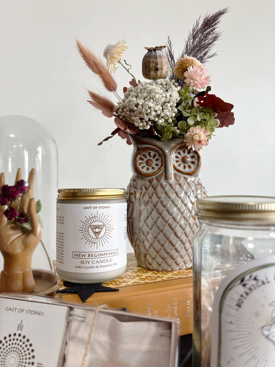 A styled tabletop scene featuring a ceramic owl-shaped vase filled with dried flowers, next to a white soy candle jar labeled Cast of Stones' New Beginnings, with other decorative jars and a glass cloche in the background.