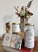 A stylized photo of a grey ceramic owl container with dried florals, next to a white candle, a glass jar Cocktail Mix, and a box of crystals and stones on top of a stack of books. In the background of the photo is a glass cloche with a wooden hand inside, holding a dried rose.