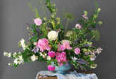 A showy, springy floral arrangement made of peonies, mock orange, veronica, and greenery in a blue distressed compote, set against a dark grey background. 