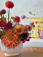 Photo of a floral arrangement made of brightly colored dahlias and cosmos in a white, ribbed vessel, sat on a wooden table. A yellow wicker couch with a throw pillow can be seen in the background.