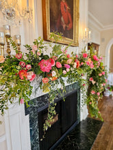 A lush floral garland with peach, coral, pink, yellow, and white flowers drapes across a mantle and fireplace. A portrait is hung above, half out of frame, and two glass candelabras hang on the wall. 