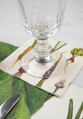 Clear stemmed napkin atop a cocktail napkin with various root vegetables, on a green and white decorated table.