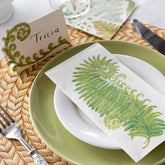 A guest napkin with a large fern illustration, sitting on a place setting made of one white plate, one green plate, and a paper placemat with a woven pattern on it. 