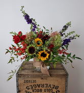 Floral arrangement in dark autumnal colors, in a glass vase wrapped with a burlap ribbon, placed on a wooden crate in front of a soft grey background. 