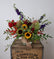 Floral arrangement in dark autumnal colors, in a glass vase wrapped with a burlap ribbon, placed on a wooden crate in front of a soft grey background. 