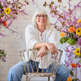 A smiling woman with white hair and glasses sits on a distressed metal chair in front of a white brick wall, surrounded by large floral arrangements in pink, yellow, and orange.