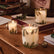 Three lit glass candles with white wax and green pine branch illustrations, arranged on a wooden table with books and holiday decor in a warm indoor setting.