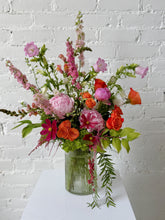 Floral Arrangement in pink, orange, and green hues set in a tall ribbed glass vase. Against a white brick background.