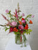 Floral Arrangement in pink, orange, and green hues set in a tall ribbed glass vase. Against a white brick background.