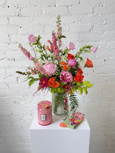 Floral Arrangement in pink, orange, and green hues set in a tall ribbed glass vase. Against a white brick background, with a pink candle and green floral socks next to the arrangement. 