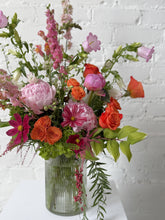 Three quarter shot of a floral arrangement in pink, orange, and green hues set in a tall ribbed glass vase. Against a white brick background.