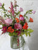 Three quarter shot of a floral arrangement in pink, orange, and green hues set in a tall ribbed glass vase. Against a white brick background.