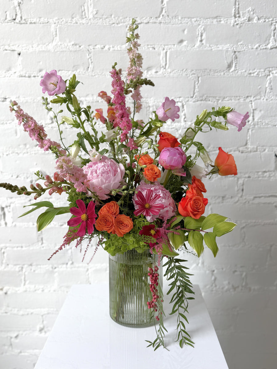 Floral Arrangement in pink, orange, and green hues set in a tall ribbed glass vase. Against a white brick background.