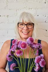A smiling woman with short white hair holds a bouquet of purple scoop scabiosa, standing against a white brick wall. 