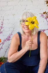 A smiling woman with short white hair sits smiling in front of a white brick wall with a yellow tulip in front of the right side of her face. She is in an all denim outfit and purple and orange flowers sit on either side of her slightly out of the frame.