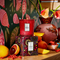 Candle sitting atop candle box, surrounded by blood oranges and other fruits, on a table with orange calla lilies. 