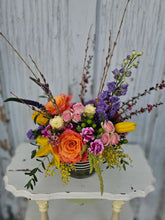 Brightly colored floral arrangement with sticks and other elements coming out of the top of it, in a vase with a black and white striped ribbon, on a white wooden table against a white background. 