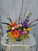 Brightly colored floral arrangement with sticks and other elements coming out of the top of it, in a vase with a black and white striped ribbon, on a white wooden table against a white background. 
