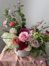 A vibrant floral arrangement featuring pink roses, white hydrangeas, burgundy carnations, and green foliage in a textured white ceramic pot, set against a soft neutral background.