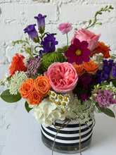 Close up of a floral arrangement in bright colors set in a glass vase with a black and white striped ribbon wrapped around it. White brick background. 