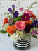 Close up of a floral arrangement in bright colors set in a glass vase with a black and white striped ribbon wrapped around it. White brick background. 