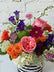 Detail shot of a floral arrangement in bright colors set in a glass vase with a black and white striped ribbon wrapped around it. White brick background. 
